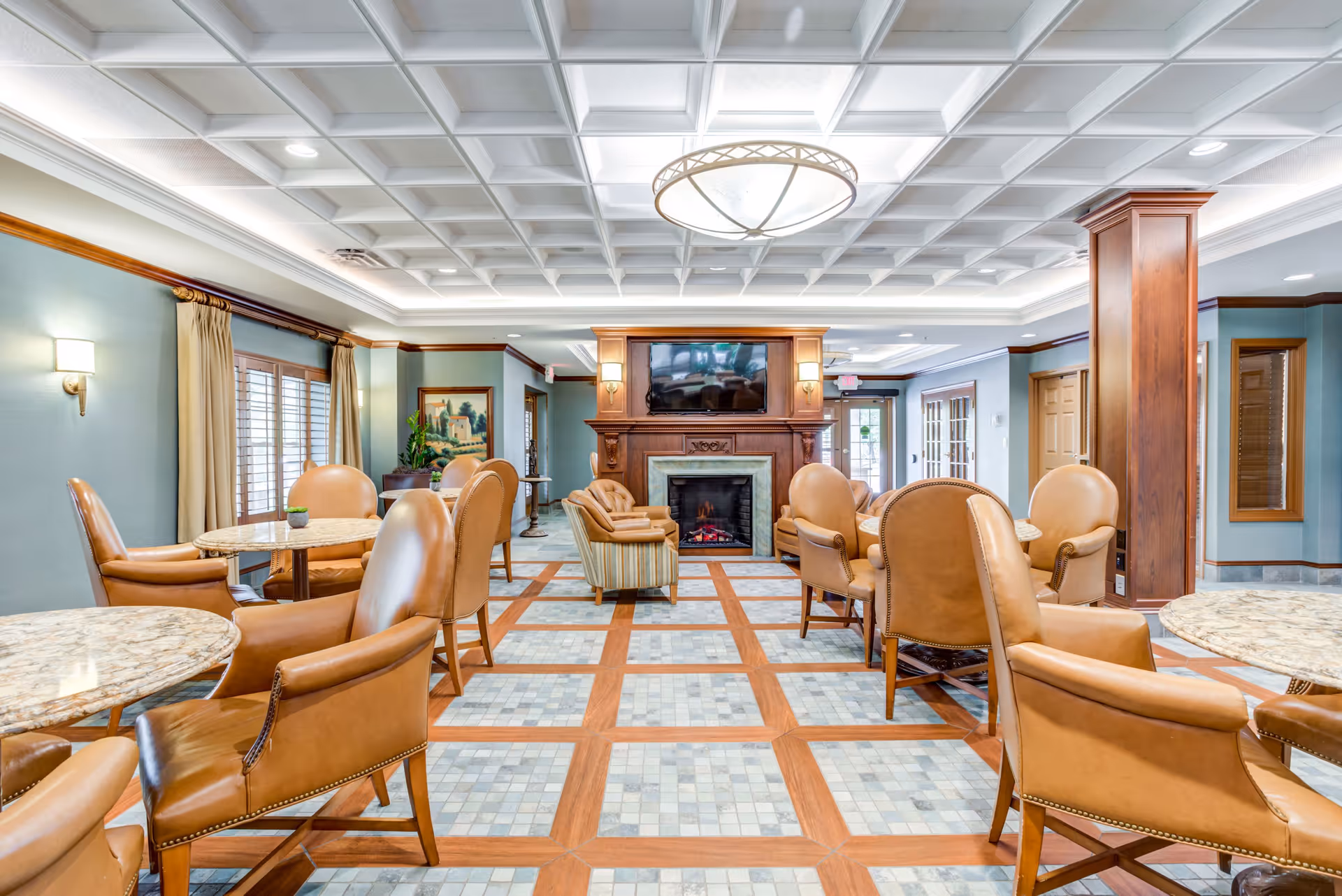 A spacious and well-lit common area with multiple round tables surrounded by tan leather chairs. The room features a decorative coffered ceiling with a large circular light fixture, a fireplace with a mounted flat-screen TV above it, and large windows with curtains allowing natural light to enter. The floor has a patterned tile design with wooden accents, and there are wooden columns and wall sconces adding to the warm ambiance.