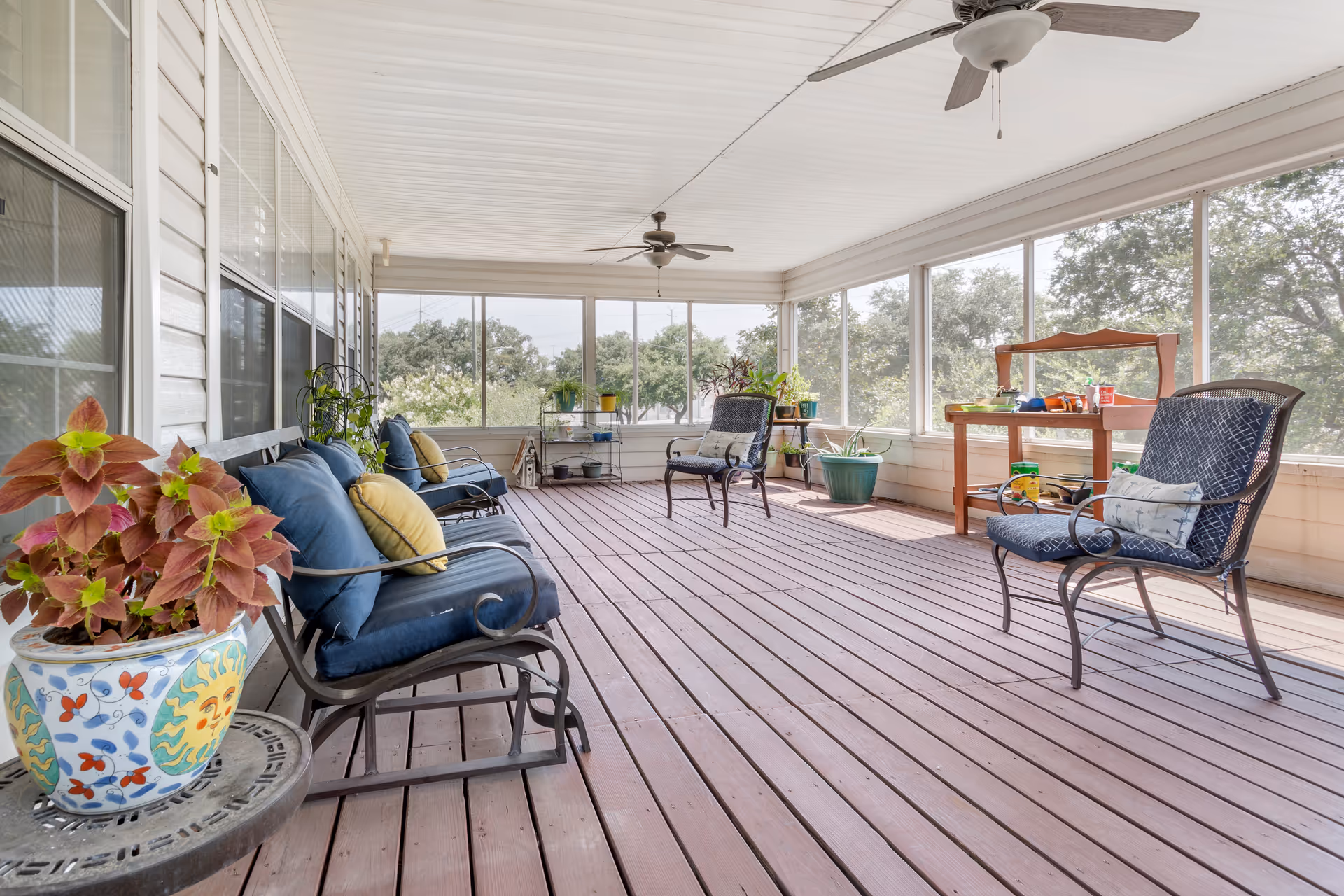 A spacious screened-in porch with wooden flooring and white ceiling. The porch is furnished with several cushioned metal chairs, some with blue cushions and yellow pillows. There are potted plants placed on a small table and along the edges of the porch. Ceiling fans are mounted on the ceiling, and large windows provide a view of trees outside.