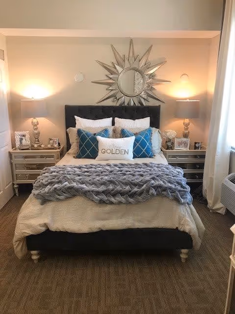 Bedroom with an upholstered bed dressed in decorative pillows and a chunky knit throw, mirrored sunburst wall decor above the headboard, and matching nightstands with lamps.