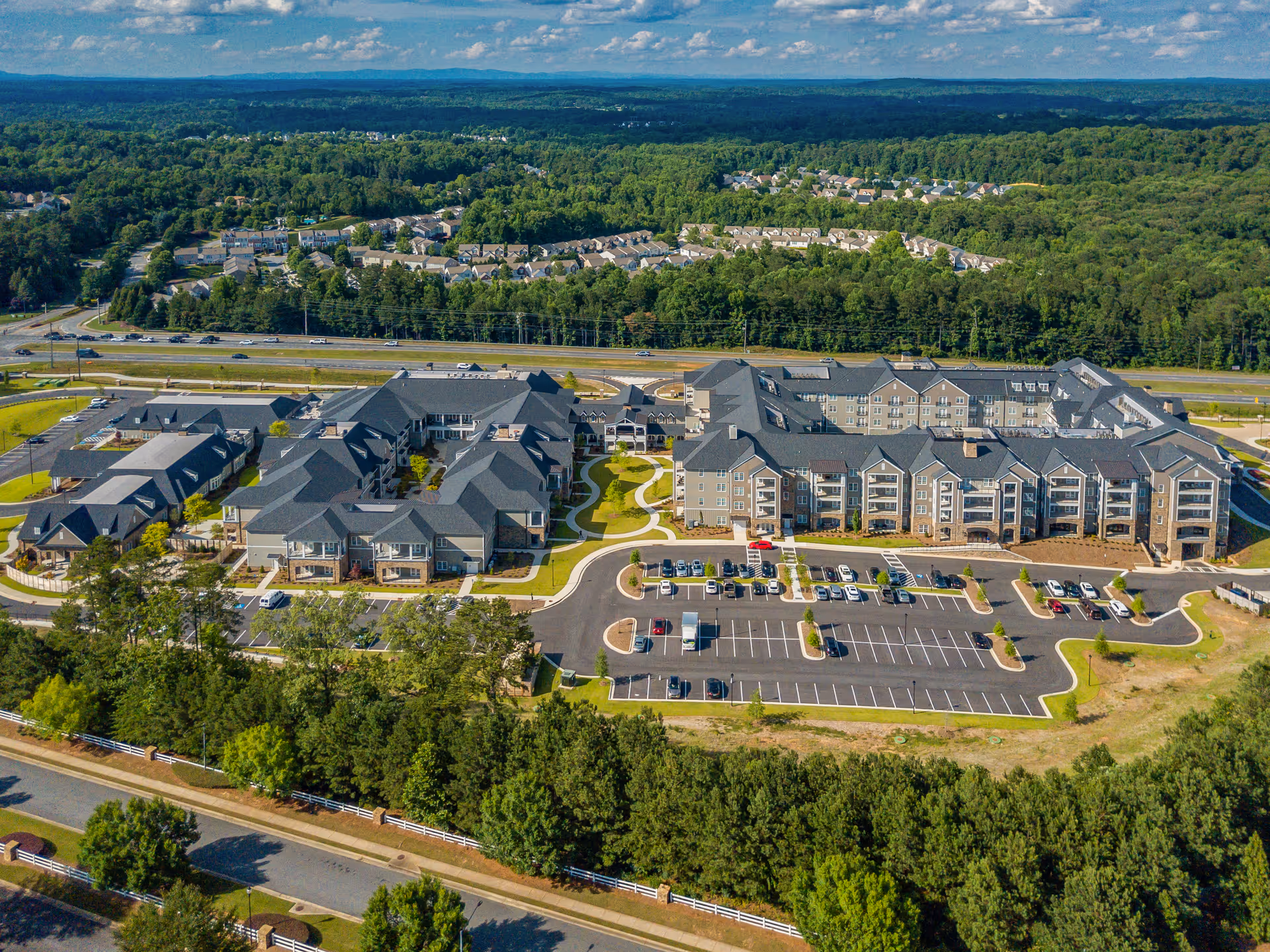 Aerial view of the Holbrook Woodstock senior living complex with multiple connected buildings, parking lot, and surrounding trees.