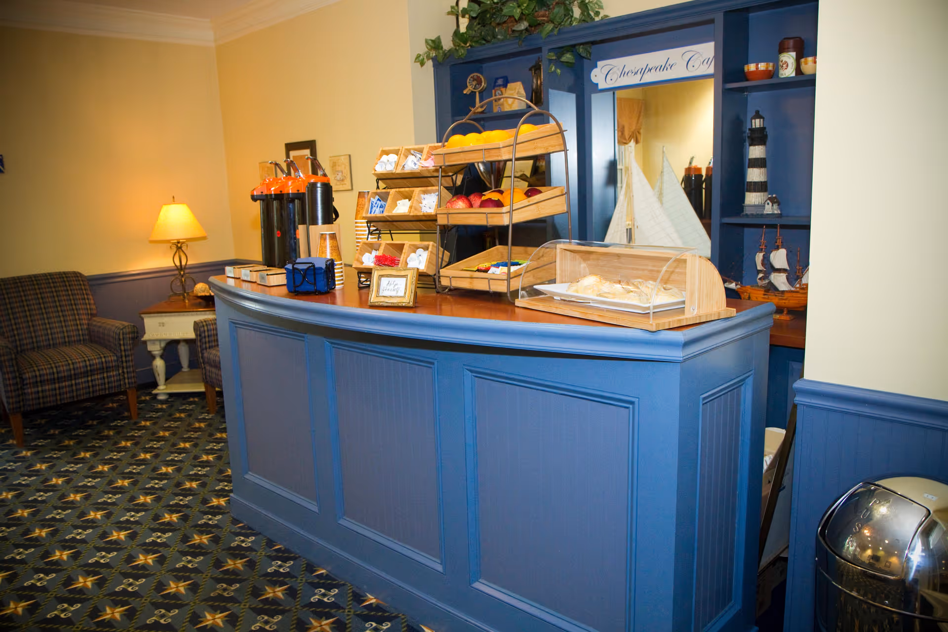 A cozy refreshment area in a senior living facility with a blue wooden counter displaying baskets of fruit, snacks, and a covered tray of pastries. Behind the counter is a blue shelving unit with nautical decorations including model ships and a lighthouse. To the left, there are two plaid armchairs and a small side table with a lamp. The carpet has a star pattern and the walls are painted in soft yellow and blue tones.