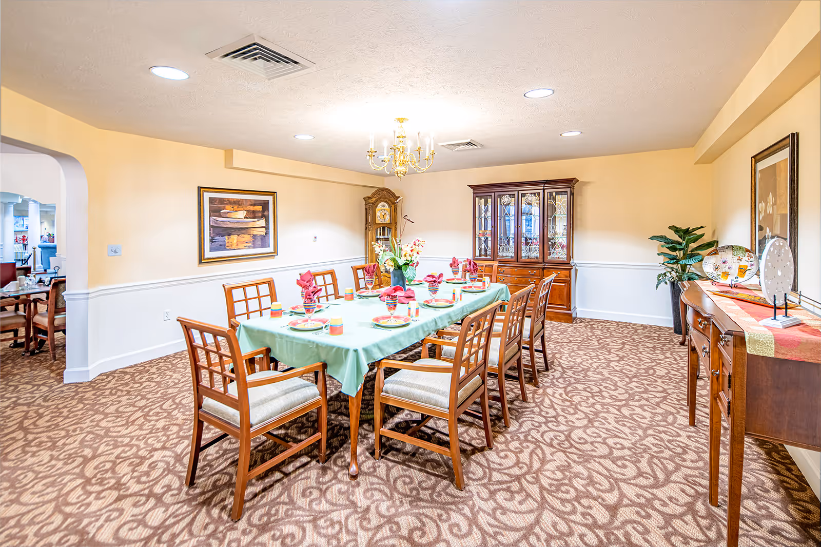 A well-lit dining room with a long rectangular table covered with a green tablecloth, set with plates, glasses, and folded red napkins. The room features wooden chairs with cushioned seats, a patterned carpet, a wooden china cabinet, a grandfather clock, framed artwork on the walls, a chandelier, and a sideboard with decorative plates and a plant.