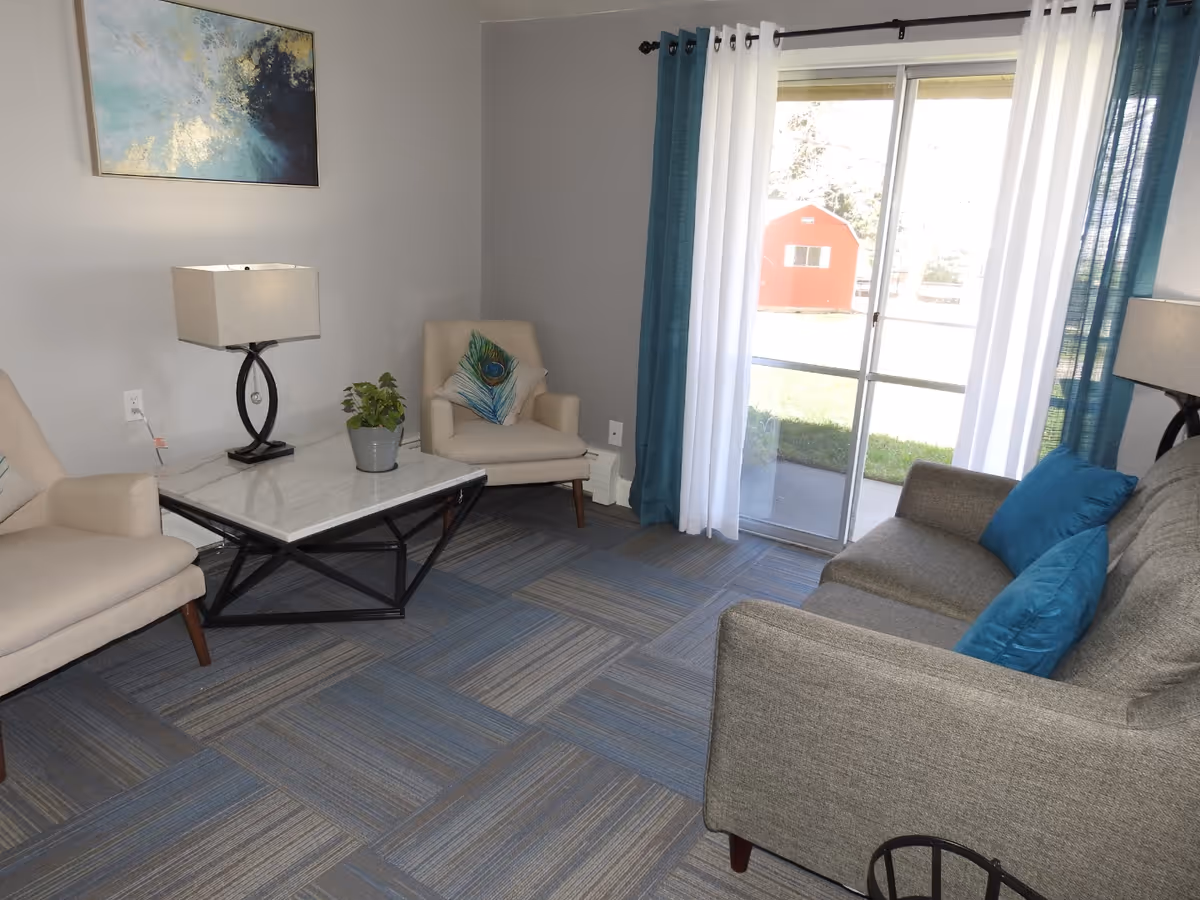 Bright living room with a sofa, two armchairs, a coffee table, lamps, and a sliding glass door with curtains.