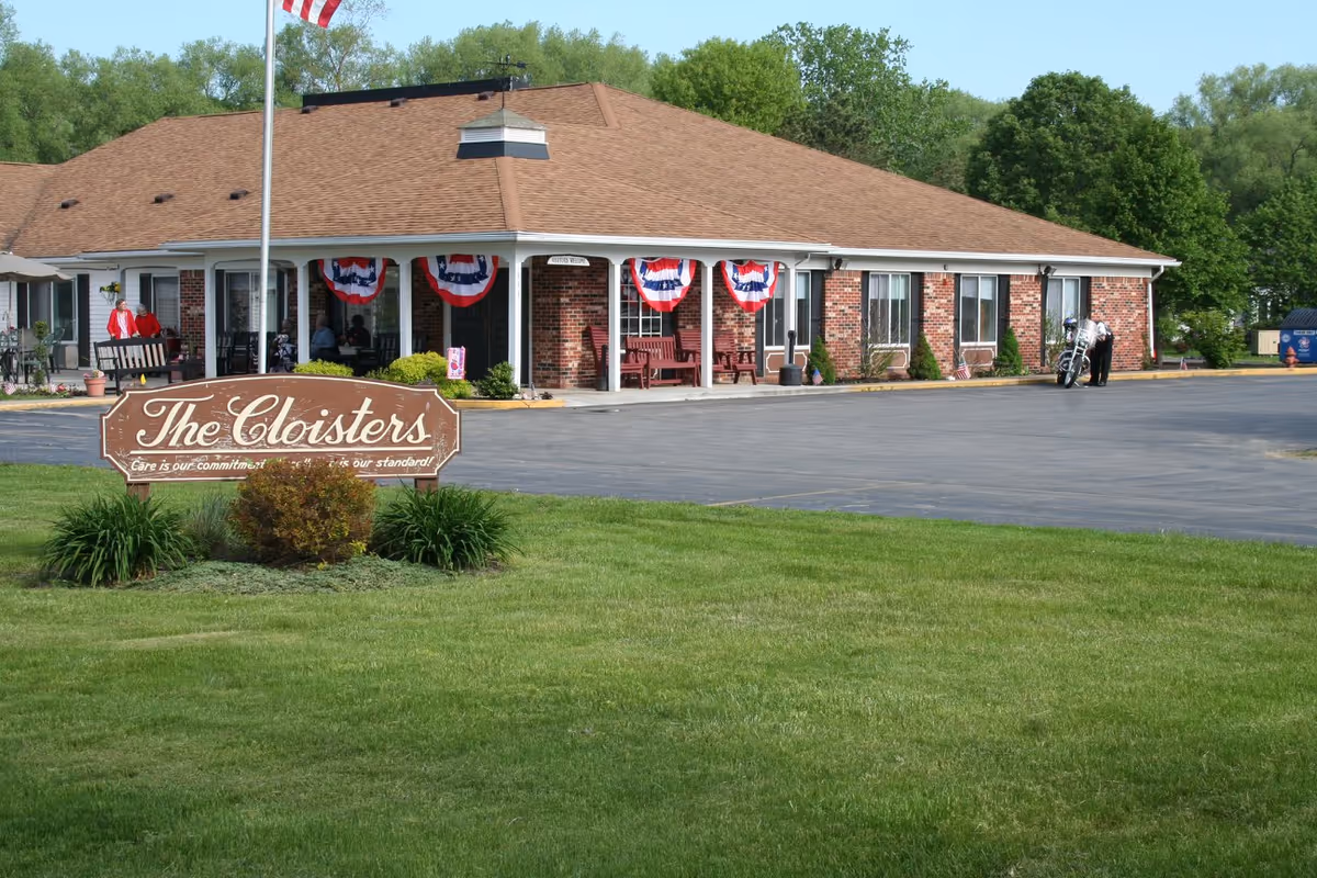 Exterior view of The Cloisters facility, a single-story brick building with a brown roof. The front porch is decorated with red, white, and blue bunting, and several people are sitting or standing near the entrance. A wooden sign in front reads 'The Cloisters' with the tagline 'Care is our commitment, quality is our standard!'. There is a green lawn in the foreground and trees in the background.