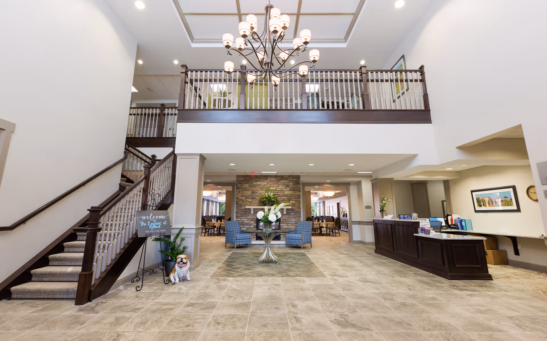 Spacious senior living facility lobby with a high ceiling, a large chandelier, a staircase with dark wood railing on the left, a reception desk on the right, and a seating area with two blue chairs and a table with flowers in the center. A welcome sign and a small dog statue are near the staircase.
