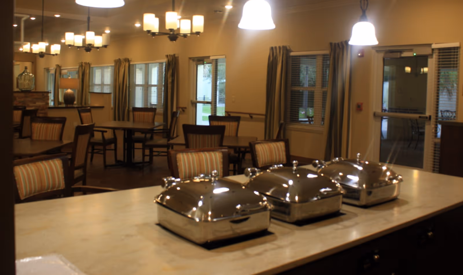 Interior view of a dining area in a senior living facility featuring multiple tables and chairs with striped cushions. In the foreground, there is a countertop with three covered stainless steel food warmers. The room is warmly lit with hanging pendant lights and chandeliers, and windows with curtains allow natural light to enter.