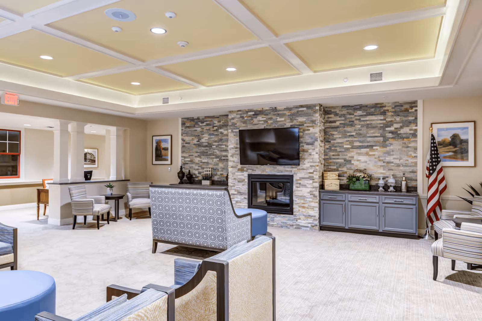 A spacious and well-lit common living area in Chantilly Heights Memory Care featuring comfortable seating with patterned and striped chairs, a stone accent wall with a mounted flat-screen TV above a fireplace, a cabinet with decorative items, framed artwork on the walls, and an American flag in the corner.