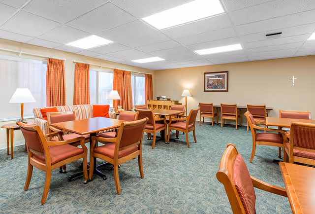 A well-lit common area with multiple wooden tables and chairs arranged for seating. The room features large windows with orange curtains, a striped sofa with orange cushions, several standing lamps, a framed picture on the wall, and a carpeted floor with a subtle pattern.