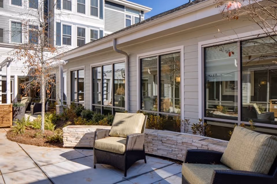 Outdoor patio area at Merrill Gardens at Woodstock featuring cushioned wicker chairs, a curved stone bench, small trees, and shrubs with large windows of the building in the background.