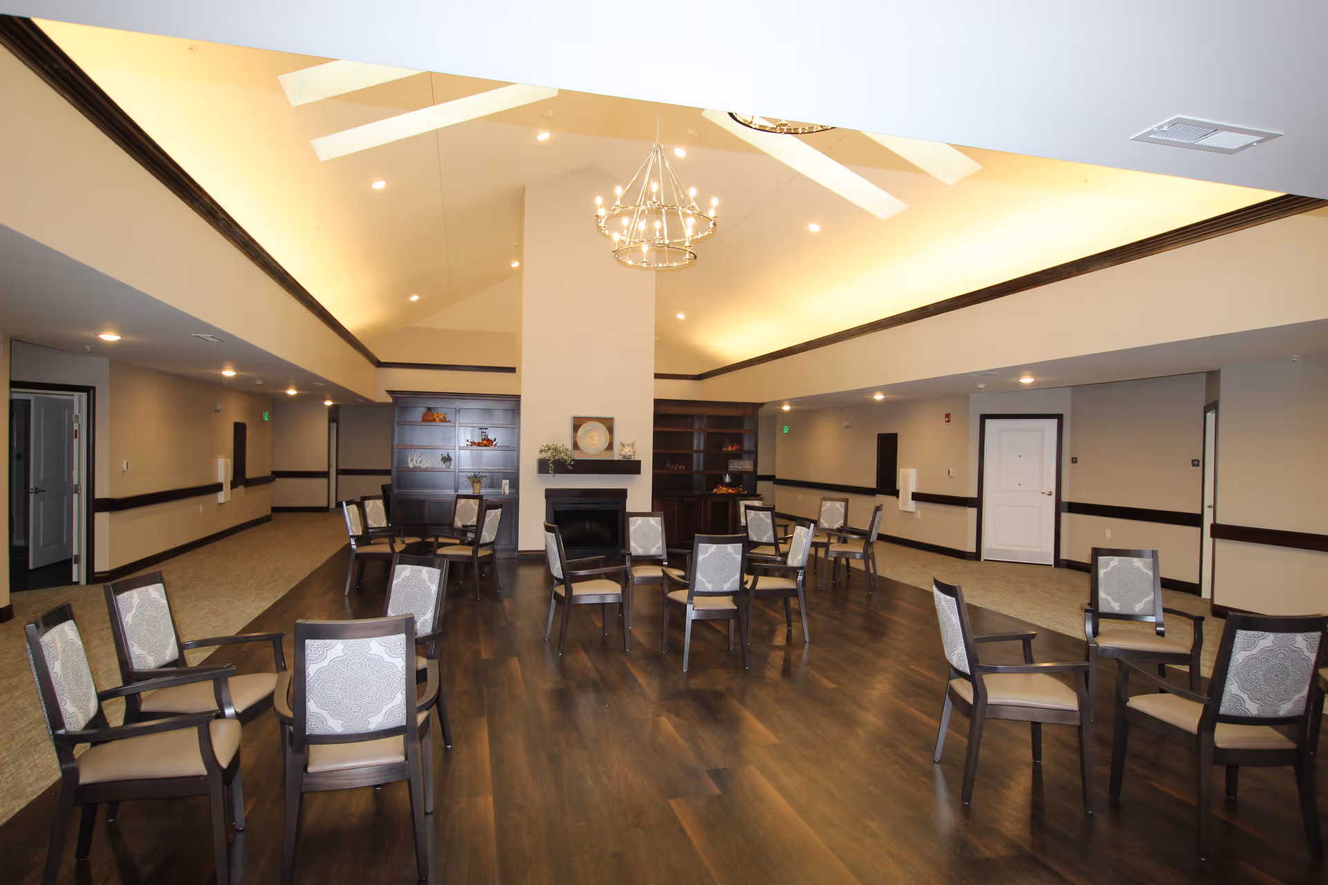 A spacious common area with multiple chairs arranged in small groups on a dark wood floor. The room features a high vaulted ceiling with a chandelier, a central fireplace, and built-in shelving units on either side of the fireplace. The walls are painted beige with dark wood trim, and there are several doors and hallways leading out of the room.