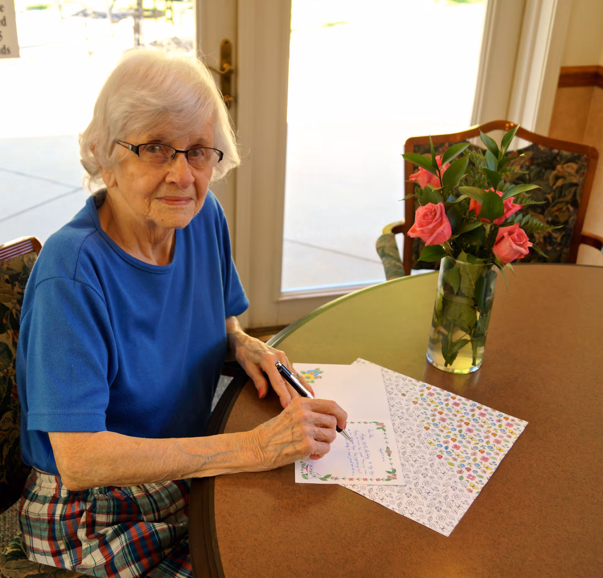 An elderly woman with white hair and glasses is sitting at a round table inside a room, writing on a floral-themed card with a pen. She is wearing a blue shirt and plaid skirt. On the table, there is a vase with pink roses and green leaves. Behind her, there is a glass door letting in natural light.