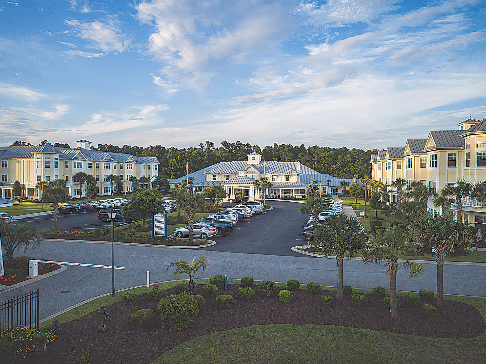 Exterior view of a senior living facility named Brightwater with multiple three-story buildings surrounding a central parking lot. The buildings have light-colored walls and metal roofs. There are palm trees and landscaped bushes around the parking area, and a clear blue sky with scattered clouds above.