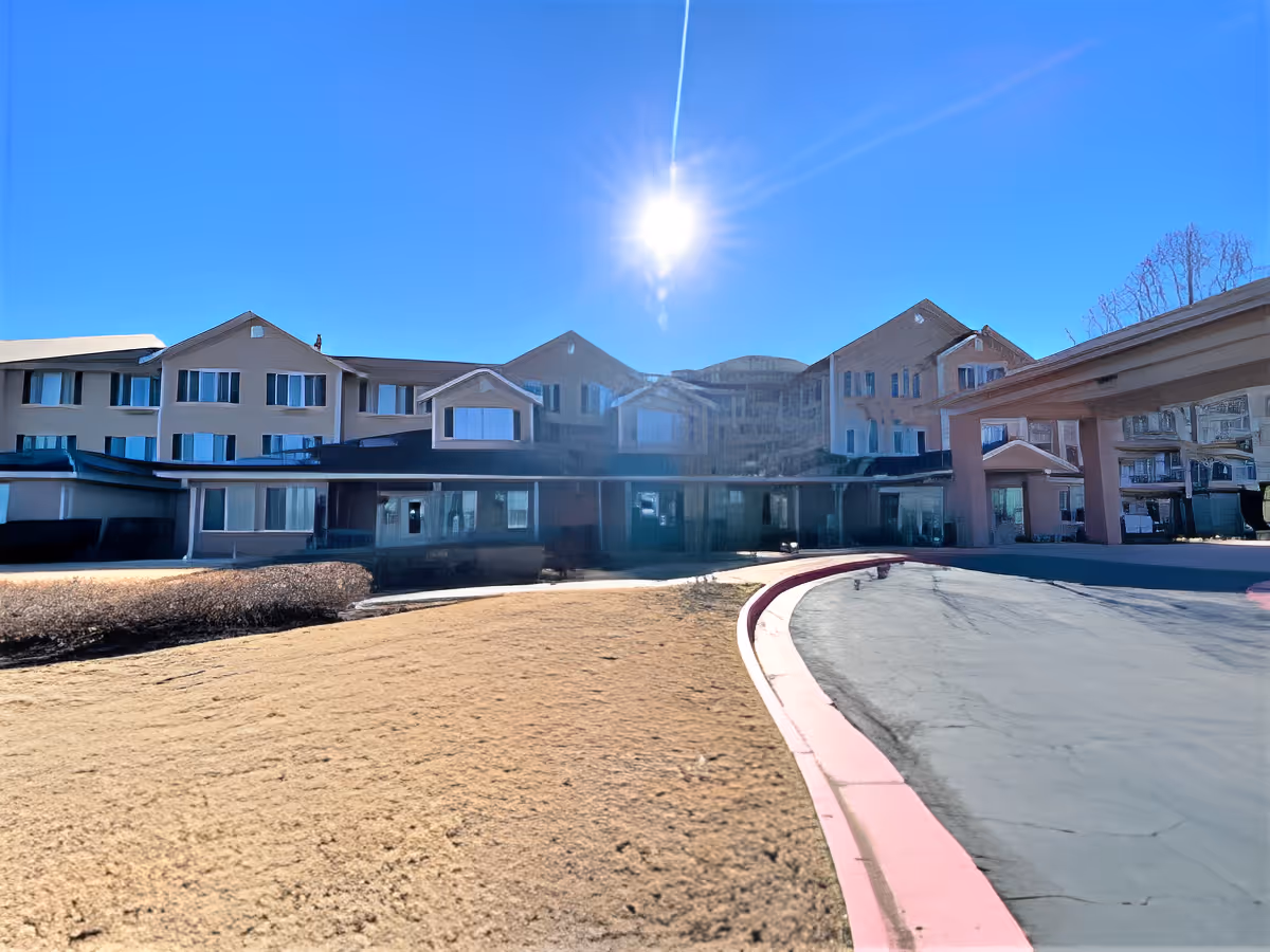 Front entrance of a multi-story senior living building with a curved driveway, lawn, and bright sun overhead.