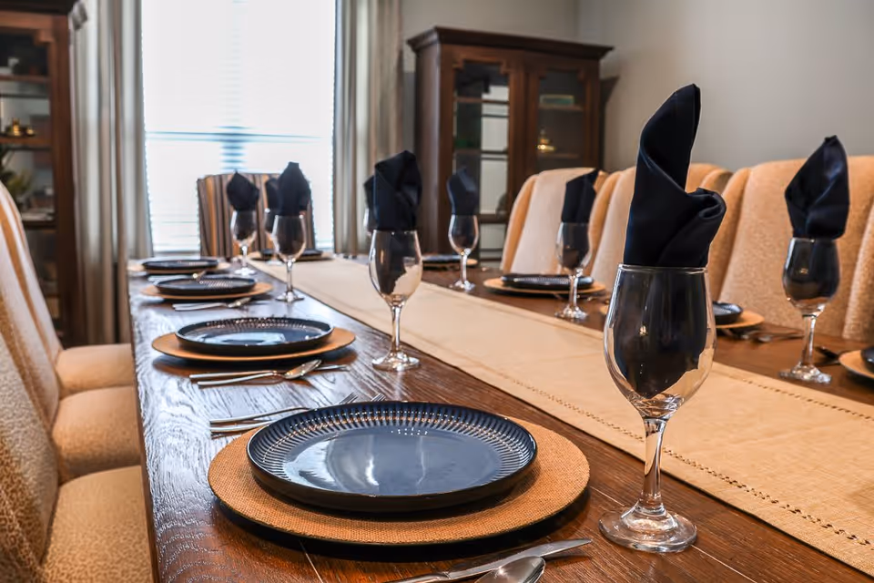 A dining table set for a meal with blue plates on woven placemats, silverware, and wine glasses with black folded napkins inside. The table is surrounded by upholstered chairs, and there is a cabinet with glass doors in the background near a window with curtains.