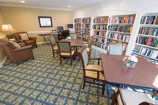 A cozy library room with patterned carpet, several wooden tables with chairs, a couple of armchairs with cushions, and built-in bookshelves filled with books along the wall. There is a small desk with two computer monitors and a printer in the corner, and a table lamp providing warm lighting.