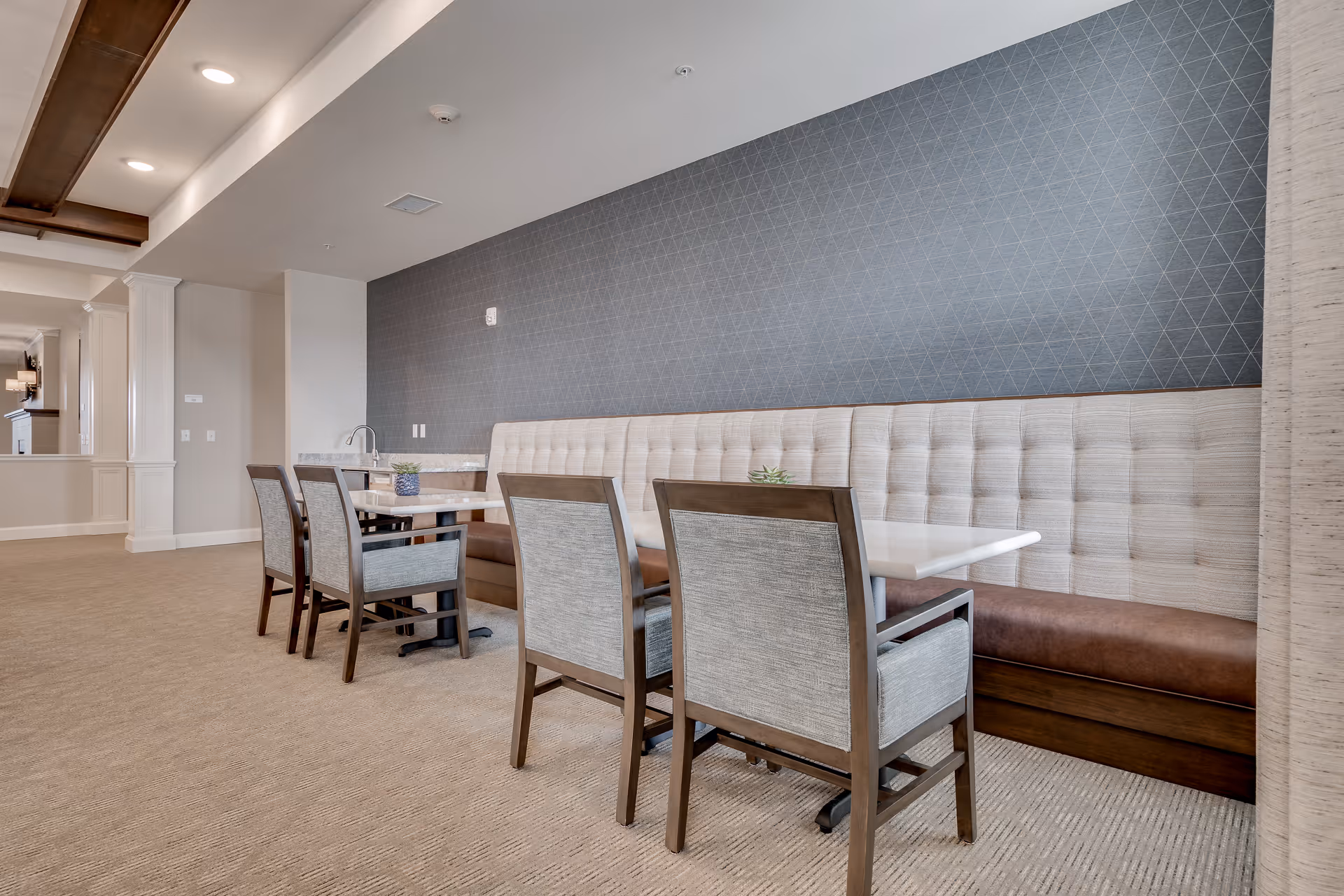 A dining area in a senior living facility featuring a long cushioned bench along a wall with geometric patterned wallpaper, paired with several tables and upholstered chairs. The space has beige carpeting and neutral-colored walls with white trim.