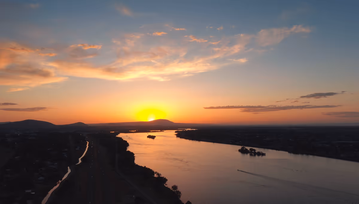 A scenic view of a river at sunset with the sun setting behind distant hills, casting an orange glow across the sky and water. There are small islands in the river and a boat creating a wake on the water. The surrounding land includes a road and scattered buildings.