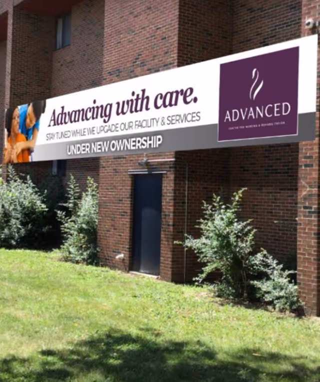 Exterior view of a brick building with a large banner that reads 'Advancing with care. Stay tuned while we upgrade our facility & services. Under new ownership.' The banner also features the logo and name 'Advanced Center for Nursing & Rehabilitation' and an image of two people smiling. There is grass and some bushes in front of the building.