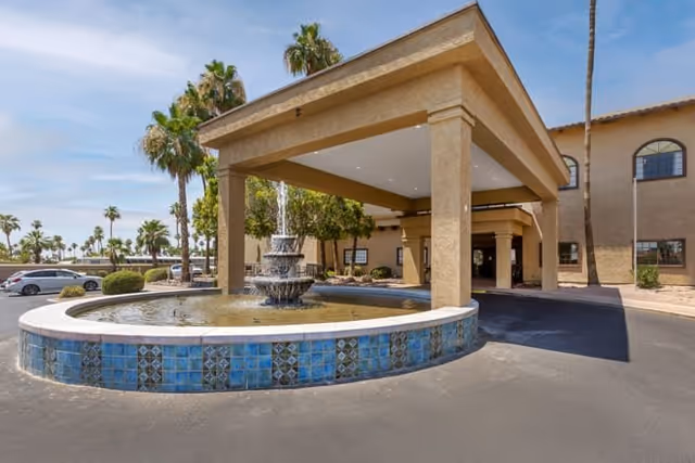 Entrance of Brookdale Springs Mesa featuring a covered drop-off area with a large circular fountain decorated with blue tiles in front. Palm trees and parked cars are visible in the background under a partly cloudy sky.