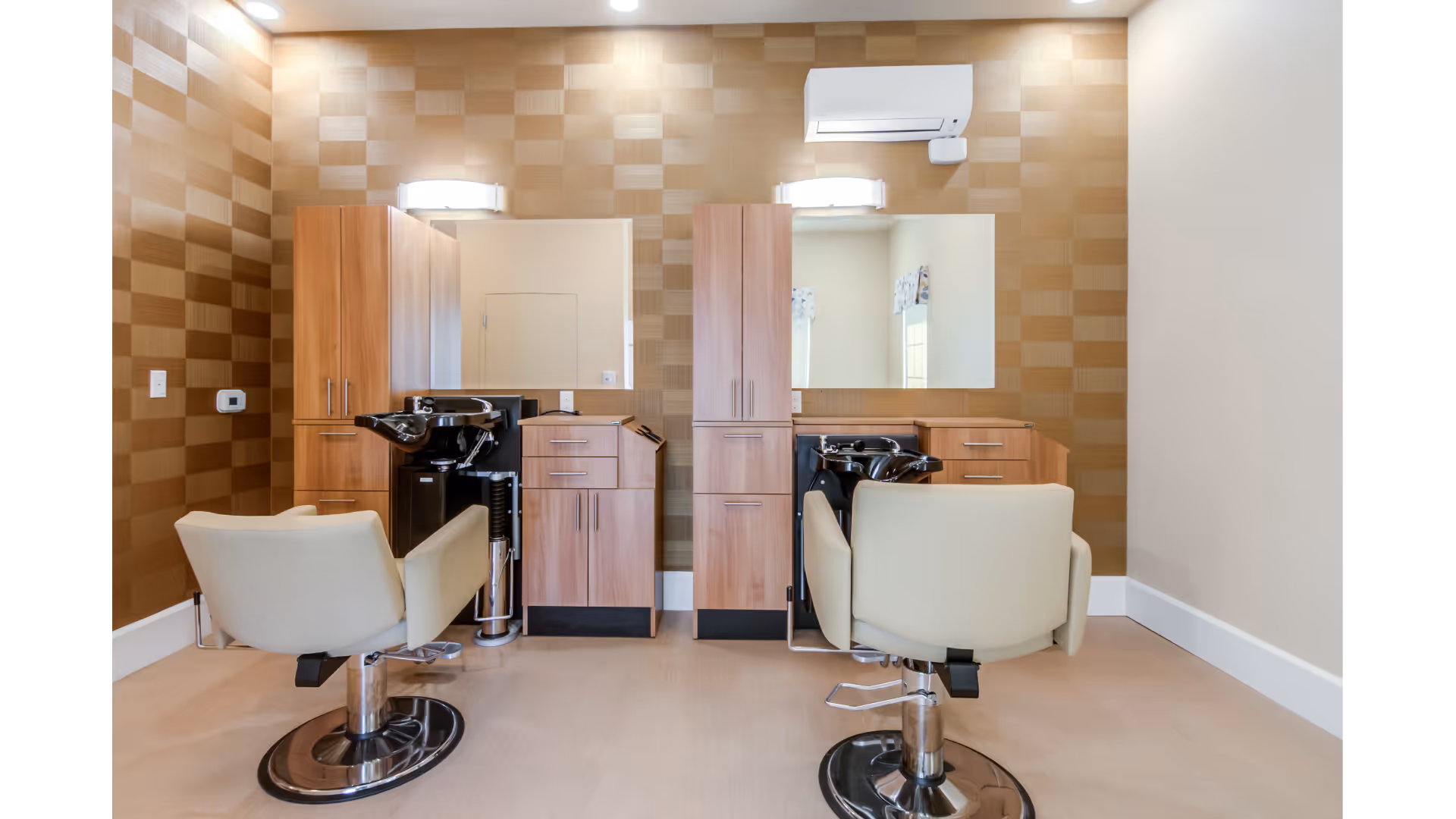 Interior view of a salon area with two beige salon chairs facing mirrors mounted on a wall with a checkered pattern. Each station has wooden cabinets and a black hair washing sink. The room is well-lit with ceiling lights and wall-mounted lights above the mirrors.