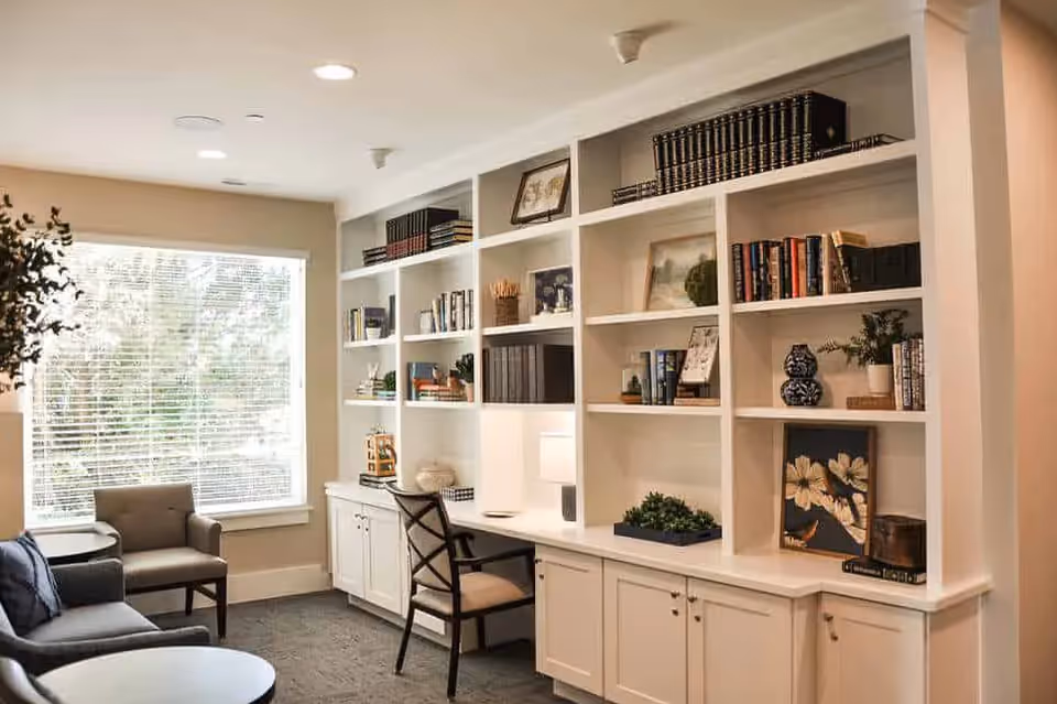 A cozy living room area with a large built-in white bookshelf filled with books, framed pictures, and decorative items. There is a desk with a chair and a small lamp integrated into the bookshelf. The room has a large window with blinds allowing natural light to brighten the space. Two armchairs and a round coffee table are positioned near the window.