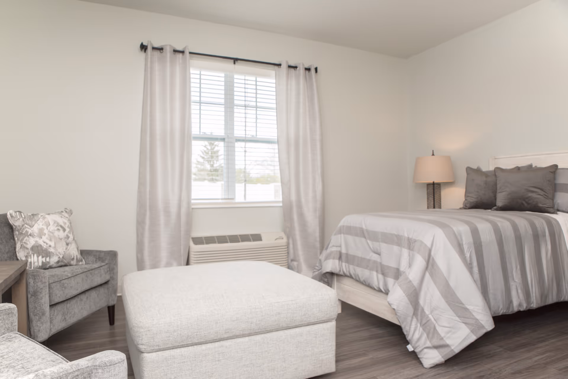 A neutral-toned bedroom with a bed covered in a striped comforter, an ottoman, an armchair, a window with curtains, and a bedside lamp.