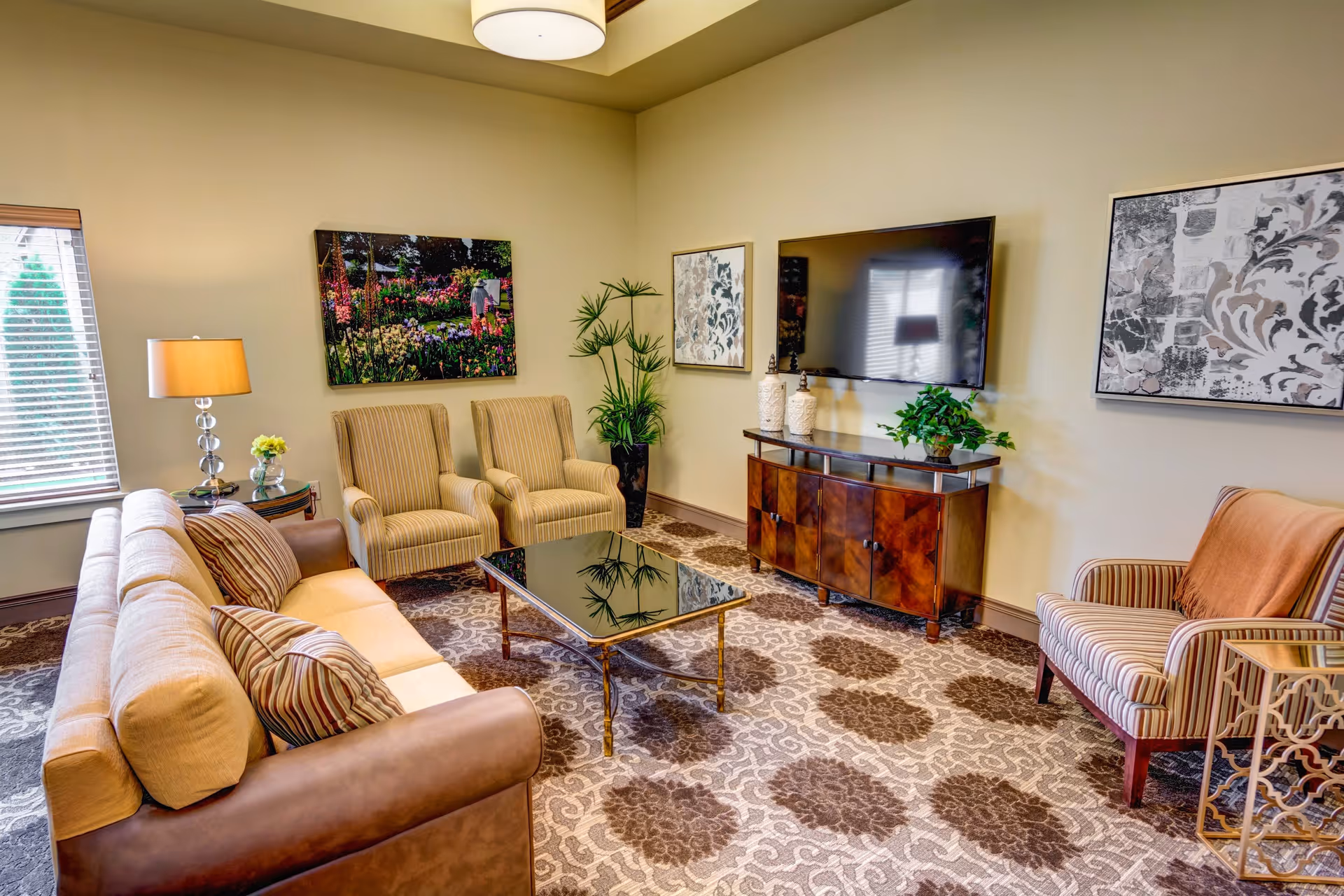 A cozy living room with a beige sofa, two striped armchairs, a glass coffee table, a wooden TV stand with a flat-screen TV mounted above it, decorative plants, and framed artwork on the walls. The room has a patterned carpet and a window with blinds letting in natural light.