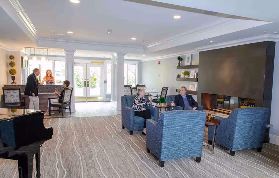 A bright and spacious senior living facility common area with a modern fireplace, blue upholstered chairs arranged around small tables, and a bar area where two people are standing and one person is seated. Large windows and glass doors allow natural light to fill the room.