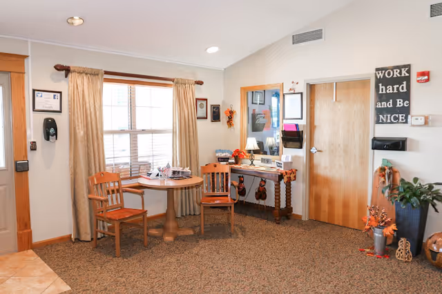 A cozy interior room with a round wooden table and two wooden chairs placed near a window with beige curtains. There is a small wooden console table against the wall with a lamp, decorations, and framed certificates or pictures above it. A wooden door is visible on the right side, and a sign on the wall reads 'WORK hard and Be NICE'. The room has a carpeted floor and neutral-colored walls with some autumn-themed decorations.