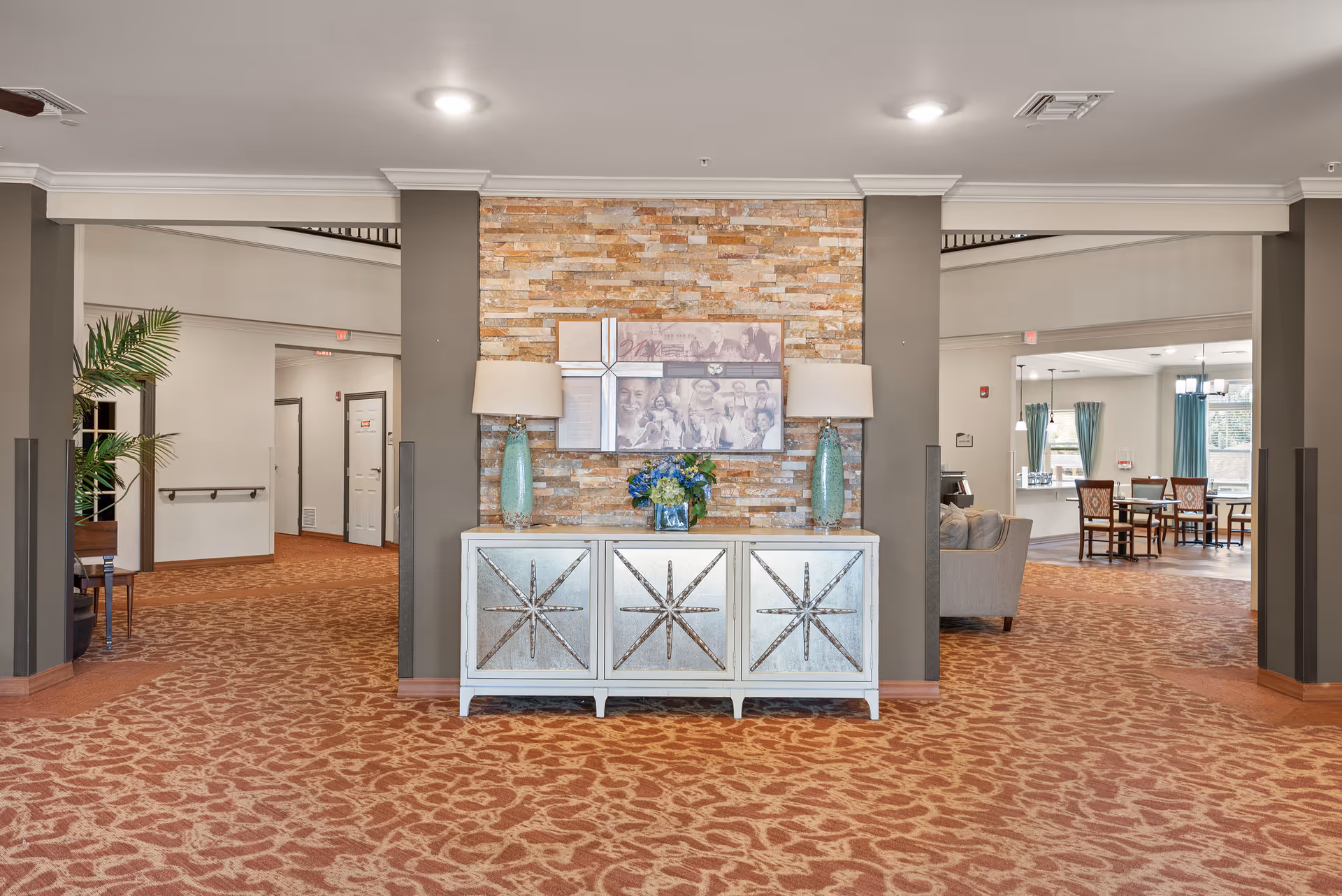 Well-lit senior living facility interior with a decorative console table and lamps centered against a stone accent wall and seating areas beyond.
