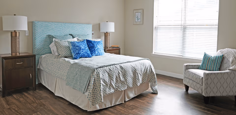 A neatly made bed with a light blue patterned headboard, multiple pillows including two bright blue ones, and a light-colored bedspread. On either side of the bed are dark wooden nightstands with matching lamps. To the right, there is a window with blinds and a cushioned armchair with a striped pillow. The room has wooden flooring and light-colored walls with a small framed picture hanging.