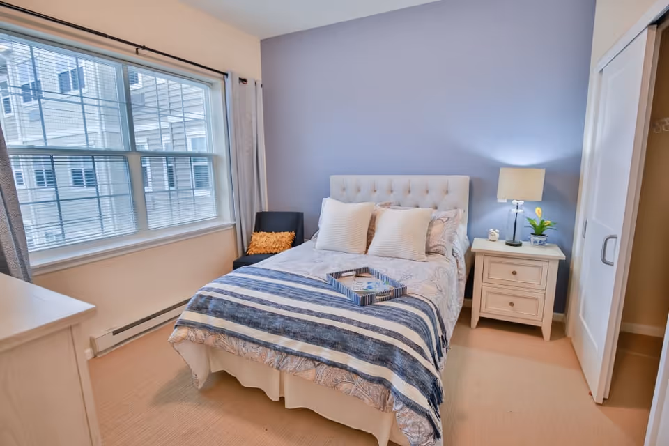 A cozy bedroom with a neatly made bed featuring a tufted headboard, two white pillows, and a blue and white striped throw blanket. There is a small tray on the bed. To the right of the bed is a white nightstand with a lamp, a small clock, and a potted plant. A black chair with an orange cushion is placed near a large window with blinds and curtains. The walls are painted light blue and beige, and there is a closet with sliding doors on the right side.