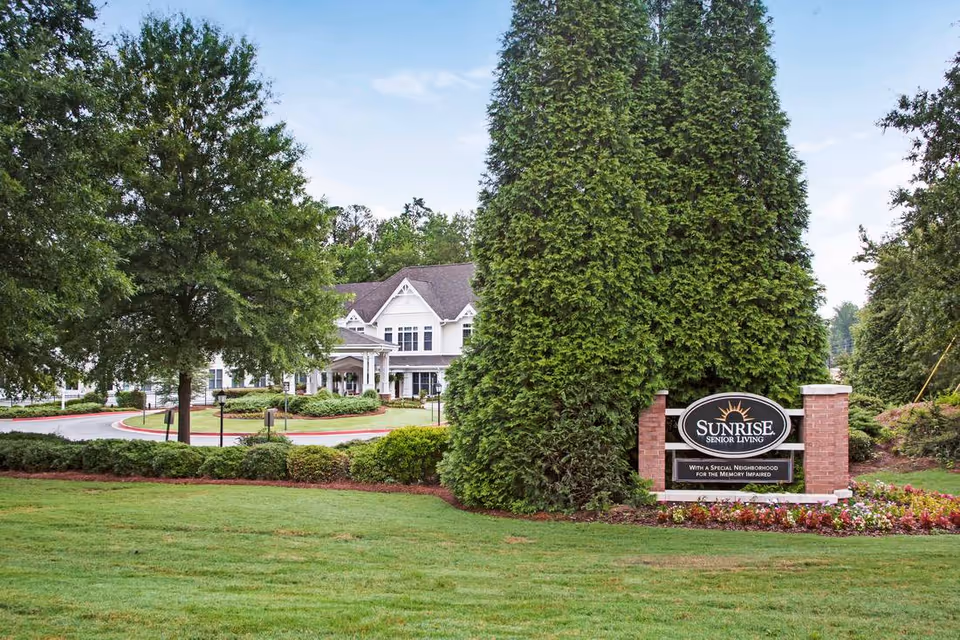Front exterior of a senior living building with a circular driveway, landscaping, and a 'Sunrise Senior Living' sign near the entrance.