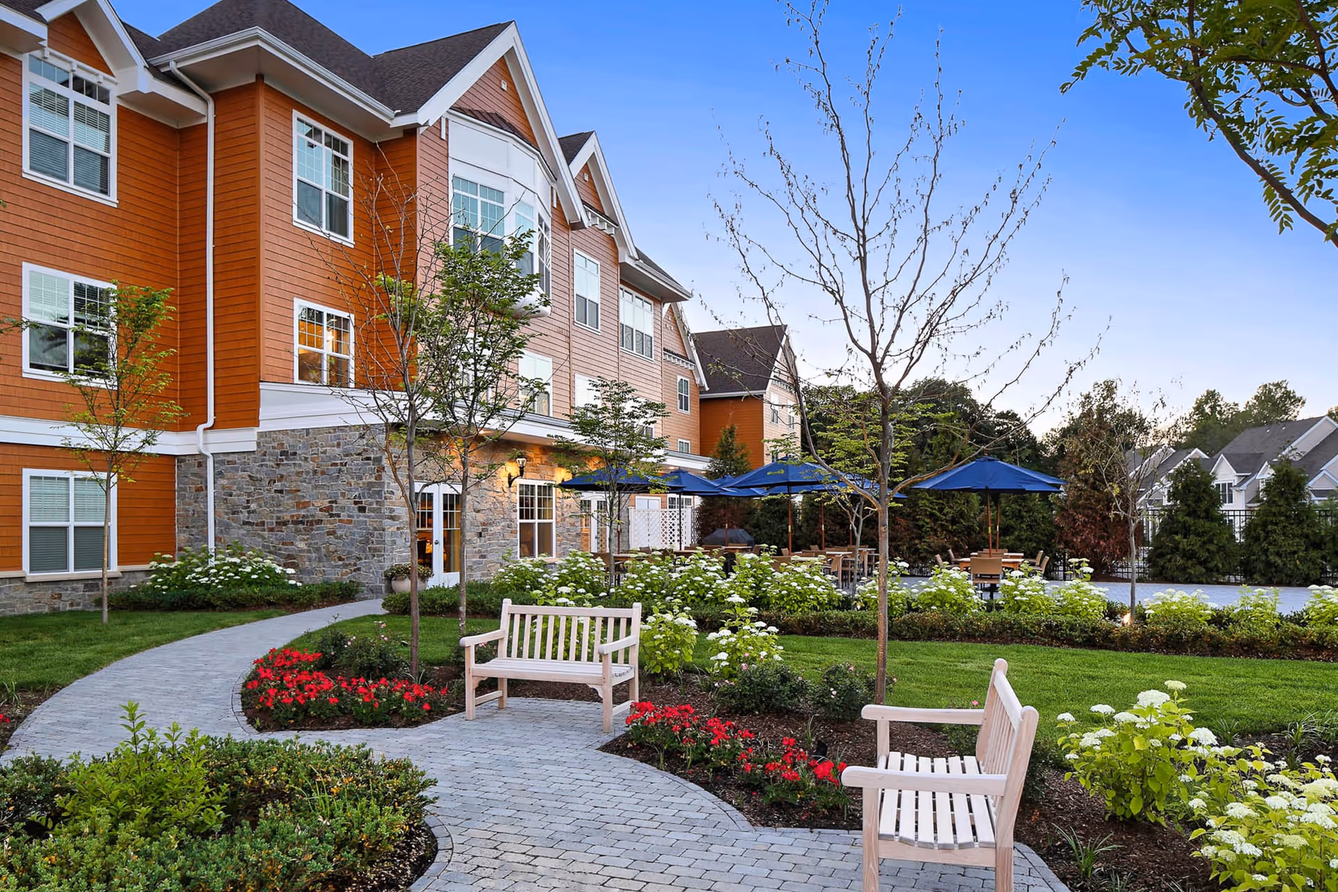 Outdoor garden area at a senior living facility with paved walkways, benches, flower beds, small trees, and tables with blue umbrellas near the building with orange siding and stone accents.