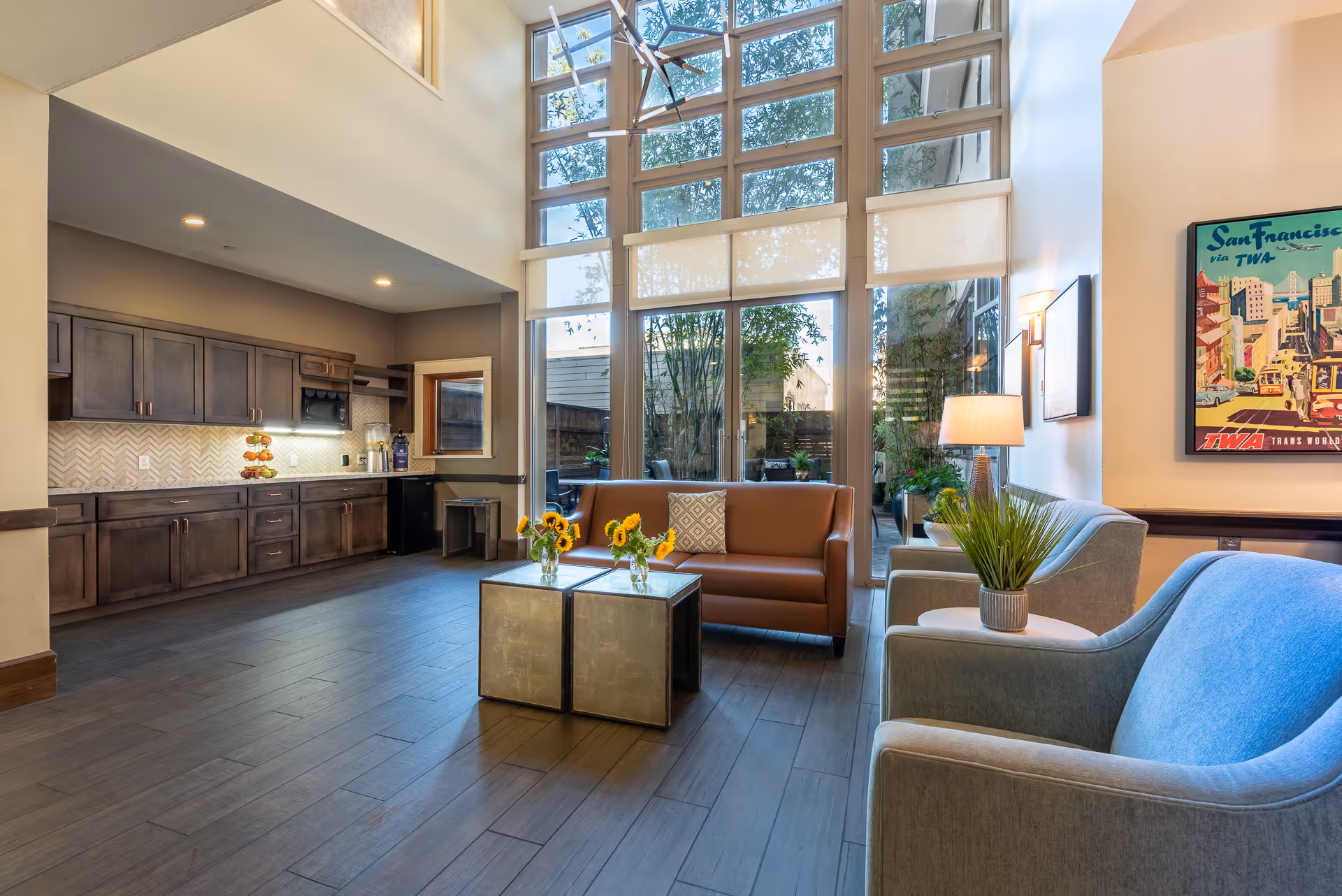 Sunlit senior living common area with tall grid windows, seating (sofa and armchairs), a coffee table with flowers, and a kitchenette along the left wall.