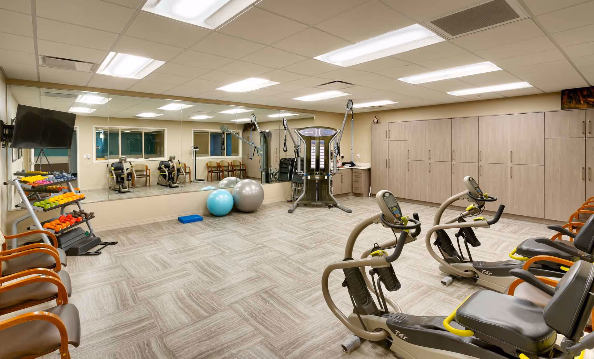 A well-lit fitness room in a senior living facility featuring exercise equipment including recumbent bikes, a multi-functional weight machine, colorful dumbbells on a rack, exercise balls, chairs lined up against the walls, large mirrors, and cabinets for storage.