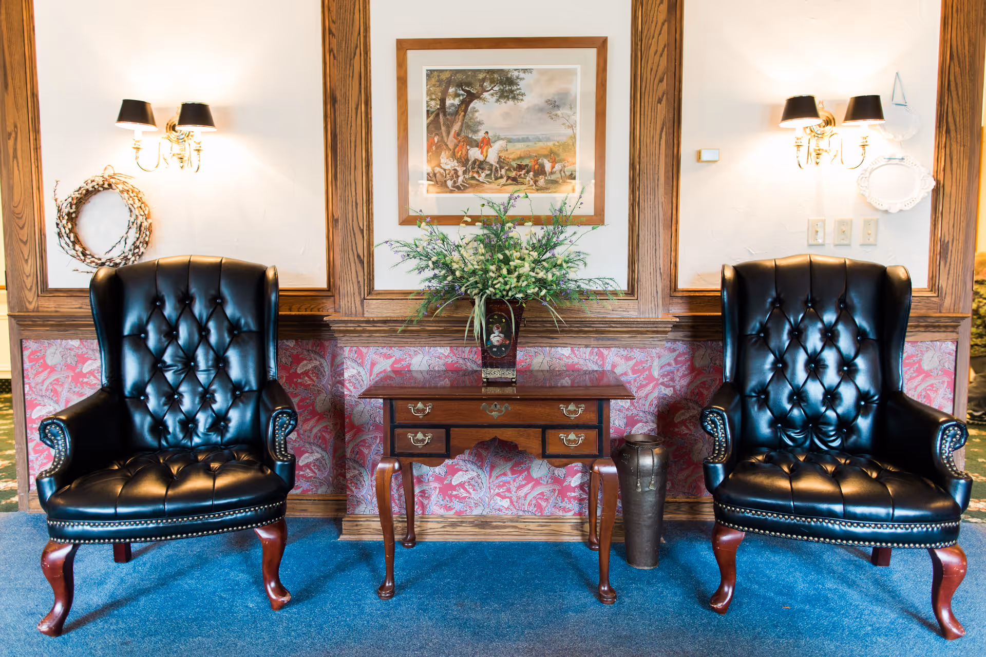 Two black leather tufted armchairs with wooden legs are positioned on either side of a wooden side table with drawers. On the table is a vase with a floral arrangement. The wall behind features wood paneling, a framed painting, and two wall-mounted lamps with black shades.