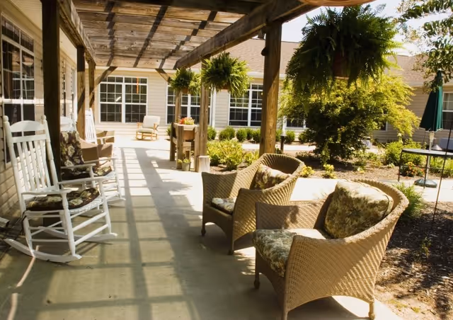 Shaded outdoor patio area with wicker chairs, rocking chairs, hanging ferns and a wooden pergola next to building windows.