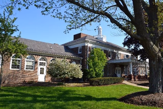 Exterior view of a brick building with white trim, surrounded by green grass, trees, and shrubs under a clear blue sky.