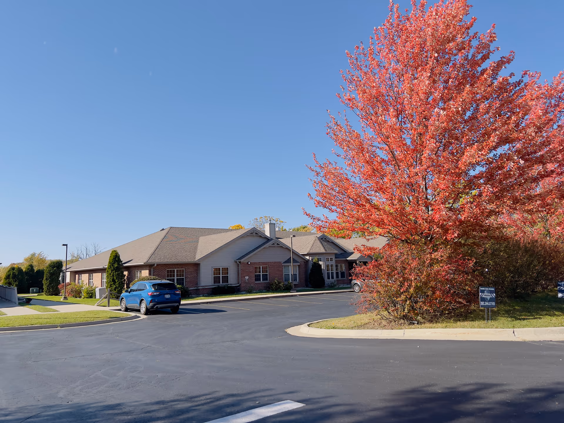Single-story brick building front with a parking lot, a blue car, and a large red-leaved tree in autumn under a clear sky.