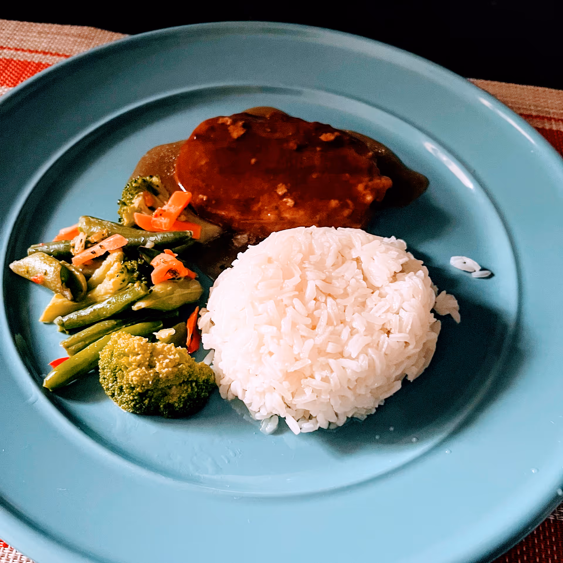 A blue plate with a serving of white rice, a piece of meat covered in brown gravy, and a side of mixed steamed vegetables including broccoli, green beans, and carrots.