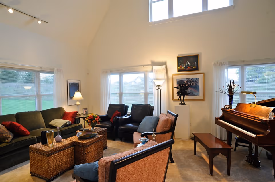 A cozy living room with a green sofa, two black leather armchairs, and two pink cushioned chairs arranged around two wicker coffee tables. The room has large windows with white curtains, a floor lamp, framed artwork on the walls, and a wooden grand piano with a bench. The space is well-lit with natural light and has a high ceiling.