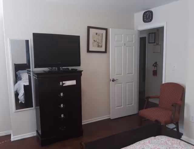 A bedroom in an assisted living facility featuring a dark wooden dresser with a flat-screen TV on top, a full-length mirror mounted on the wall, a framed picture, an open door labeled with the number 3, and a red upholstered armchair. Part of a bed with a patterned bedspread is visible in the foreground.