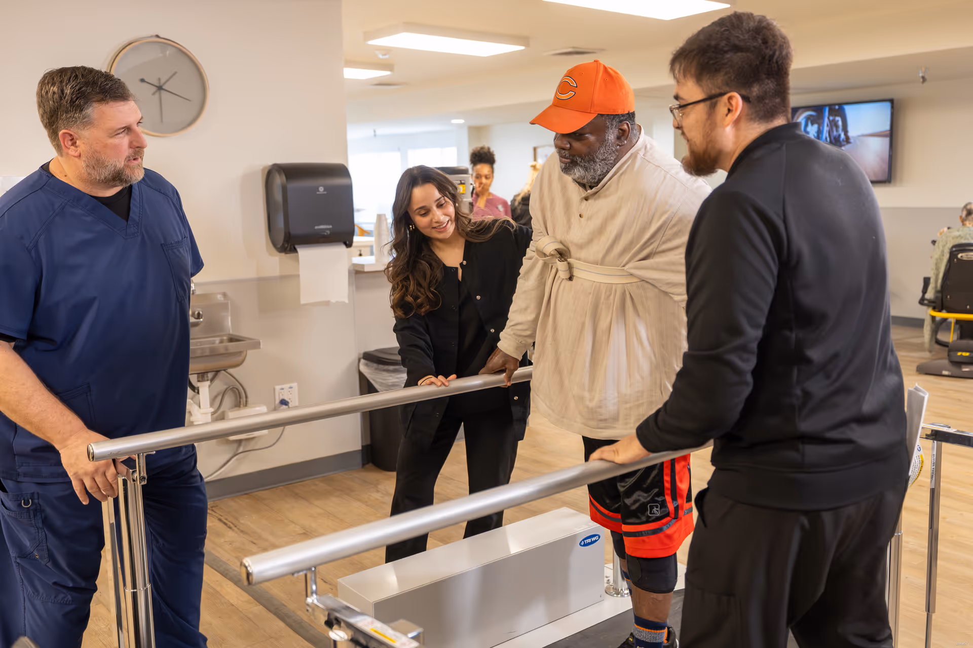 An elderly man wearing an orange cap and beige jacket is using parallel bars for physical therapy in a rehabilitation facility. He is assisted by three staff members, two men and one woman, who are attentively supporting and encouraging him. The room has a clock on the wall, a paper towel dispenser, and a television in the background.