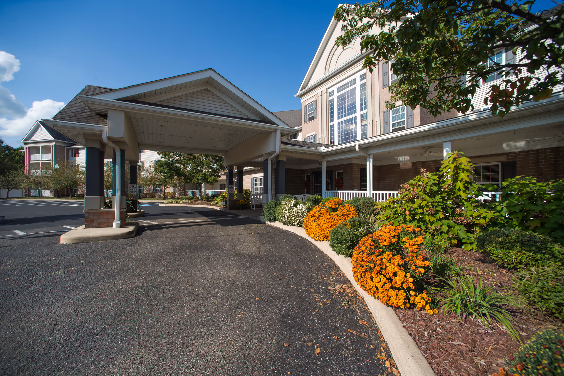 Entrance area of a senior living facility with a covered drop-off zone, landscaped flower beds with orange and white flowers, and a multi-story building with large windows in the background under a partly cloudy sky.