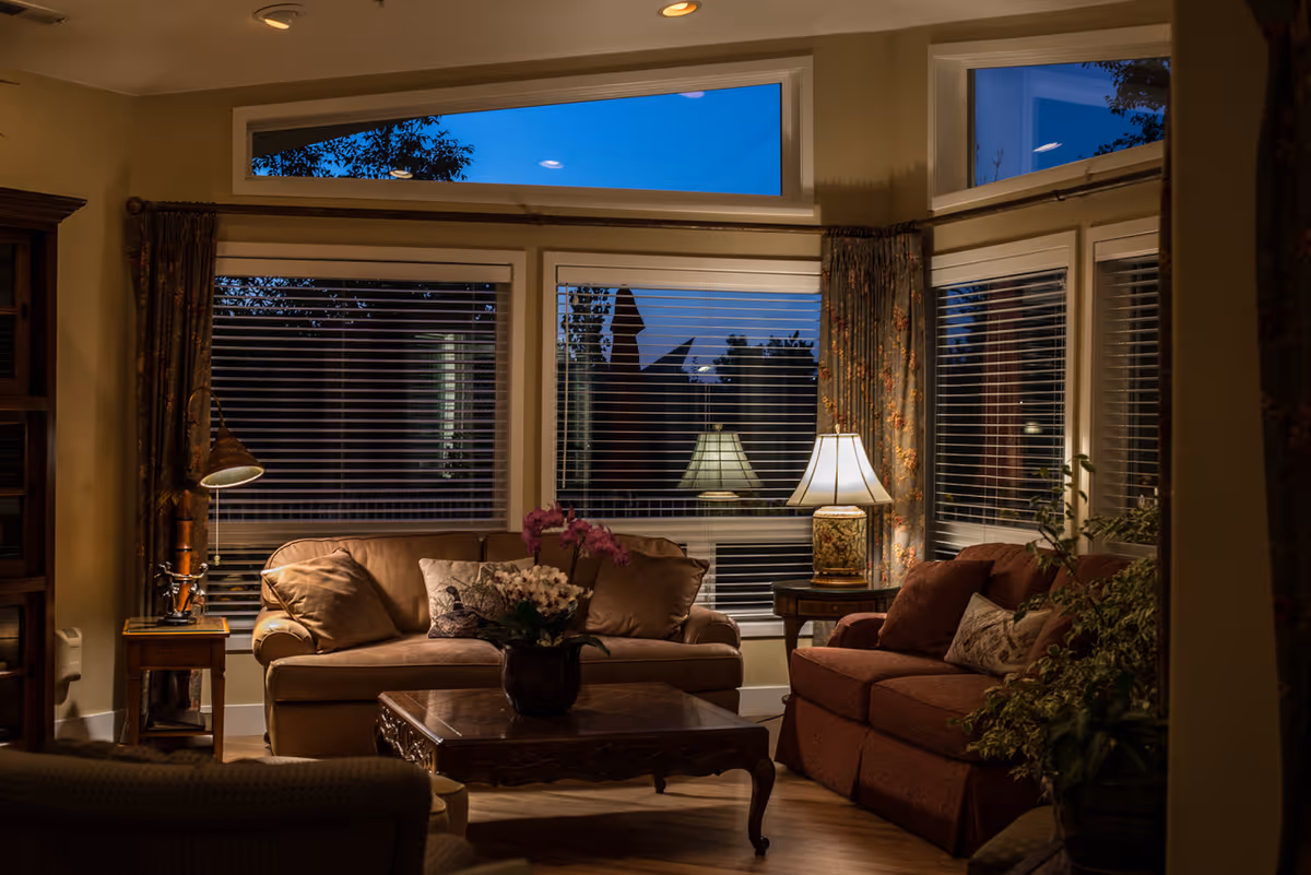 Cozy living room with two sofas, a wooden coffee table with a flower vase, two side tables with lamps, large windows with blinds and floral curtains, and warm lighting during evening time.
