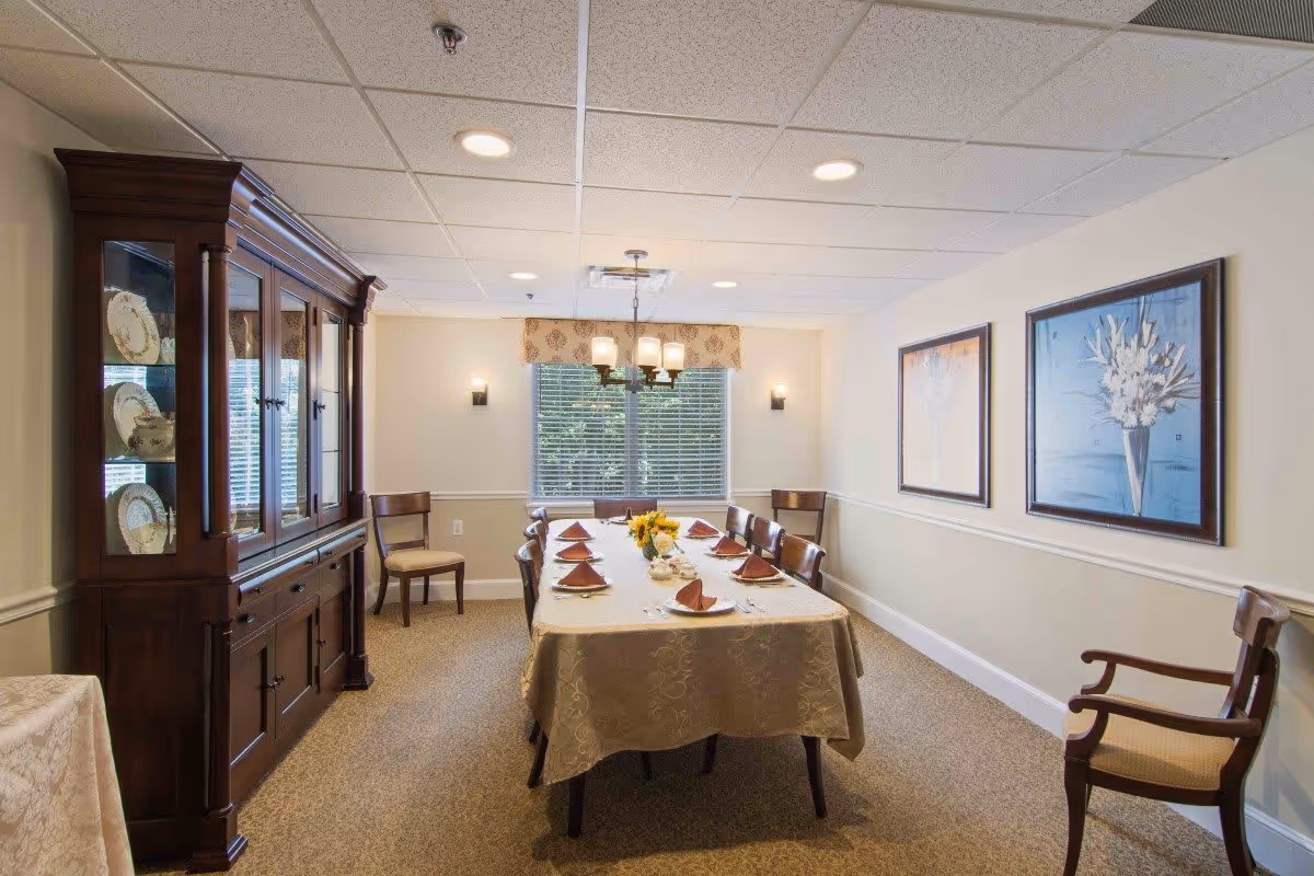 A dining room with a long rectangular table covered with a beige tablecloth, set with plates, napkins, and silverware. There are eight wooden chairs around the table and two additional chairs against the walls. A wooden china cabinet with decorative plates is on the left side. The room has beige walls with white trim, two framed floral paintings on the right wall, a window with blinds and a valance at the far end, and a ceiling light fixture above the table.