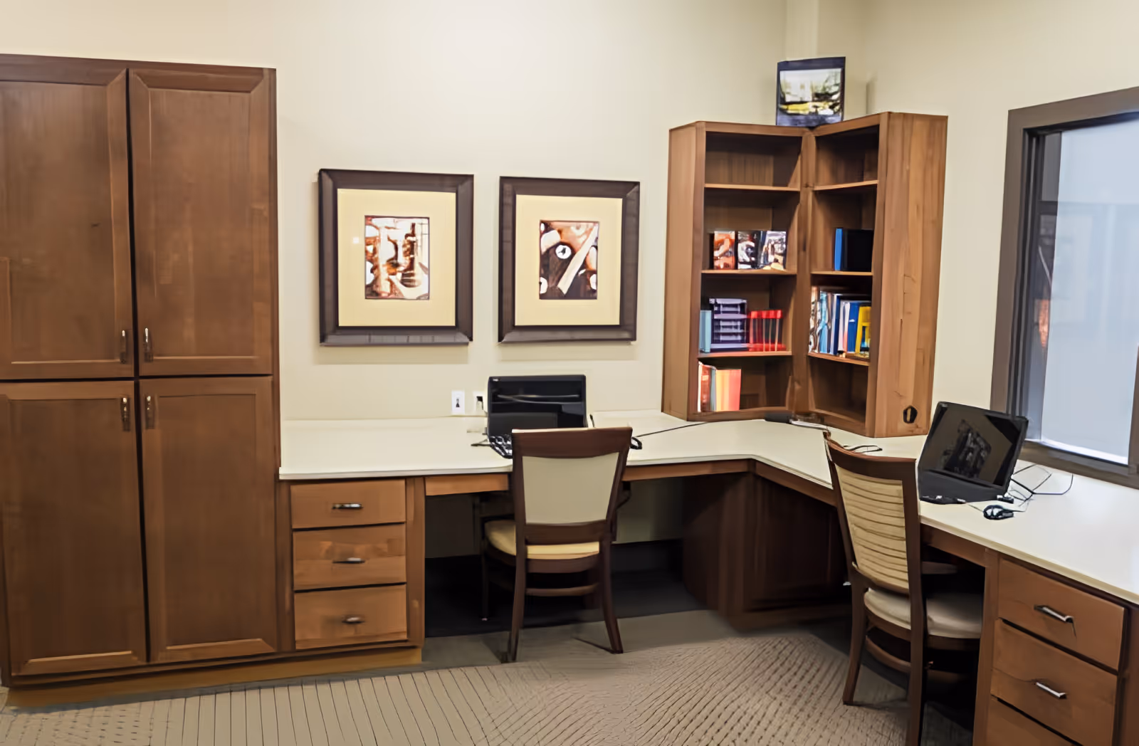 An office or study area with a corner desk featuring two chairs, two laptops, and wooden cabinets and shelves filled with books and decorative items. Two framed abstract artworks hang on the wall above the desk.