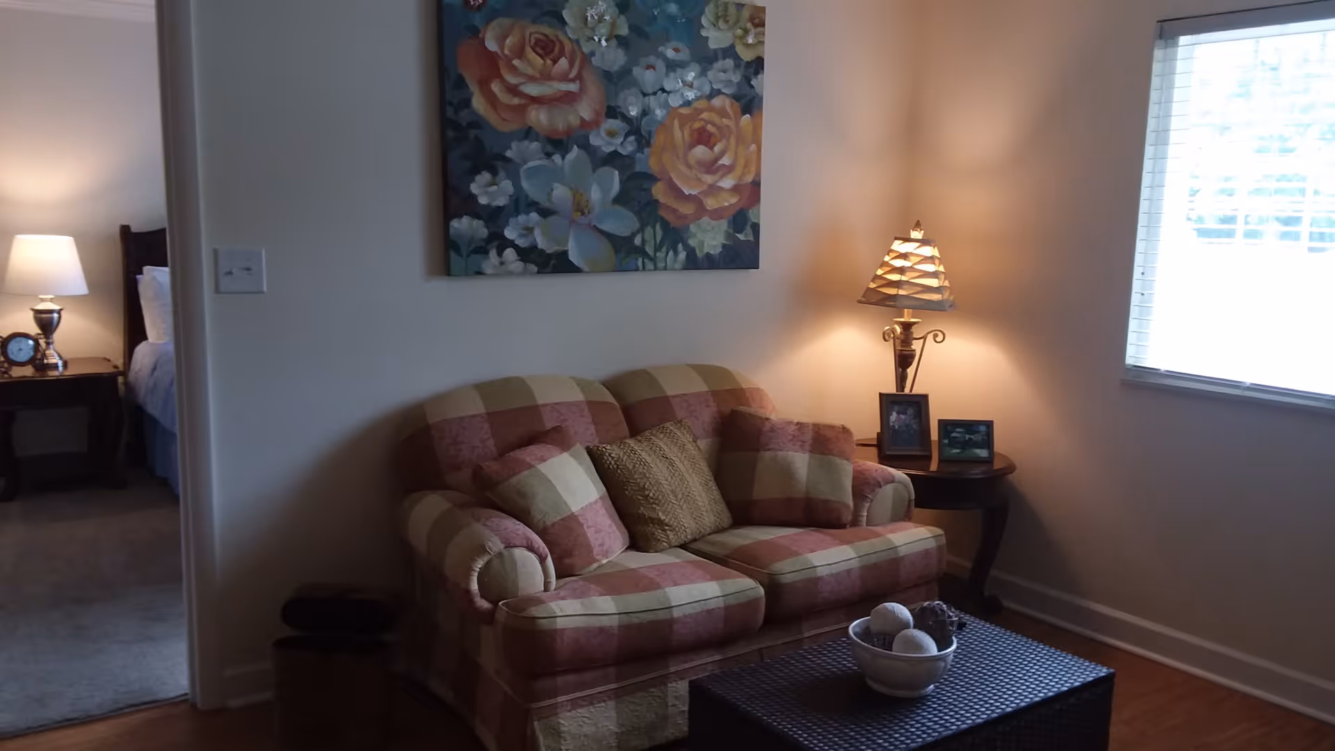 A cozy living room area with a plaid loveseat adorned with three cushions. Above the loveseat hangs a floral painting. To the right, a side table holds a lamp and two framed photos, next to a window with blinds. To the left, an open doorway reveals a bedside table with a lamp and an alarm clock in a bedroom.