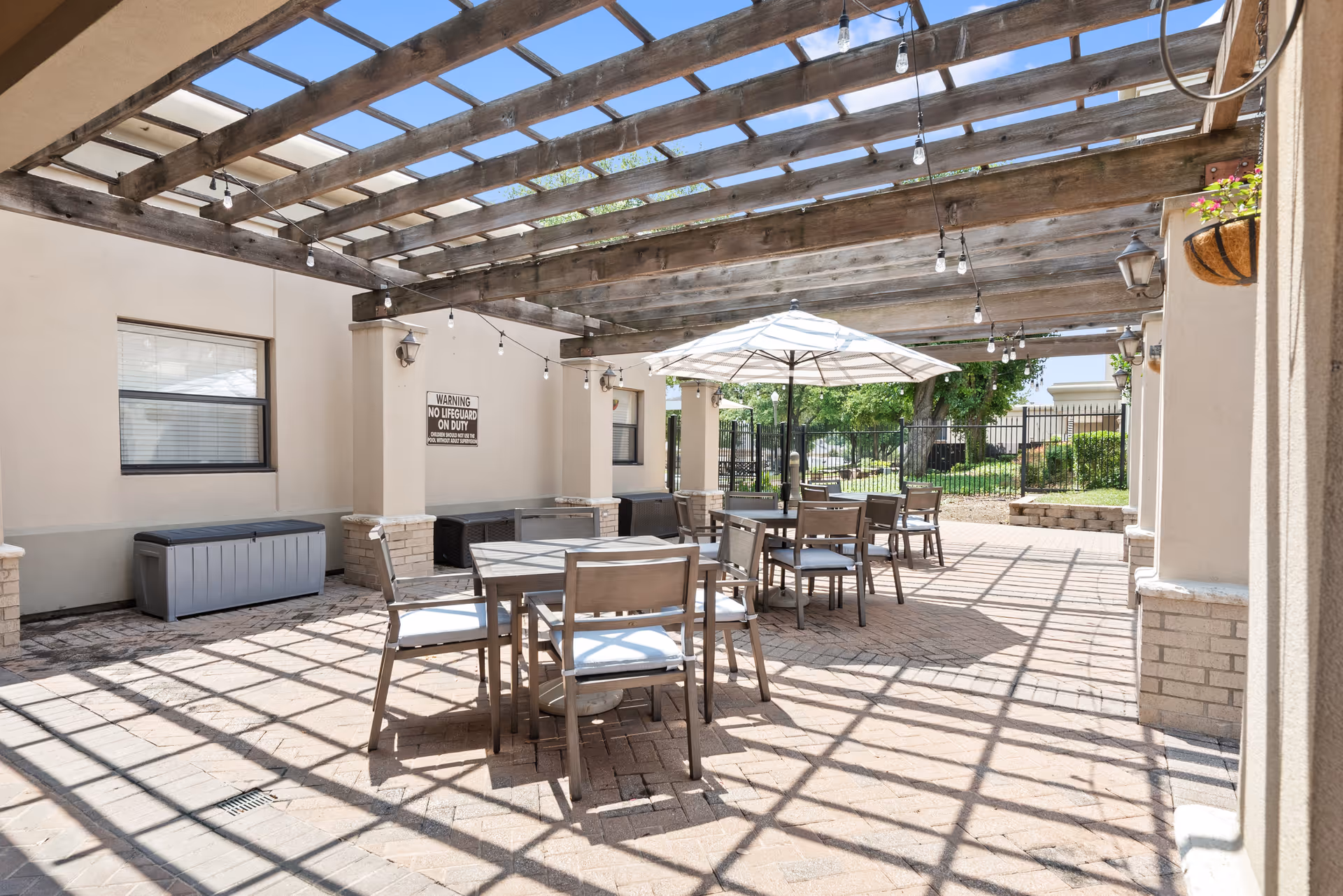Shaded outdoor patio at Parc Place with a wooden pergola, tables, chairs, and a central umbrella.